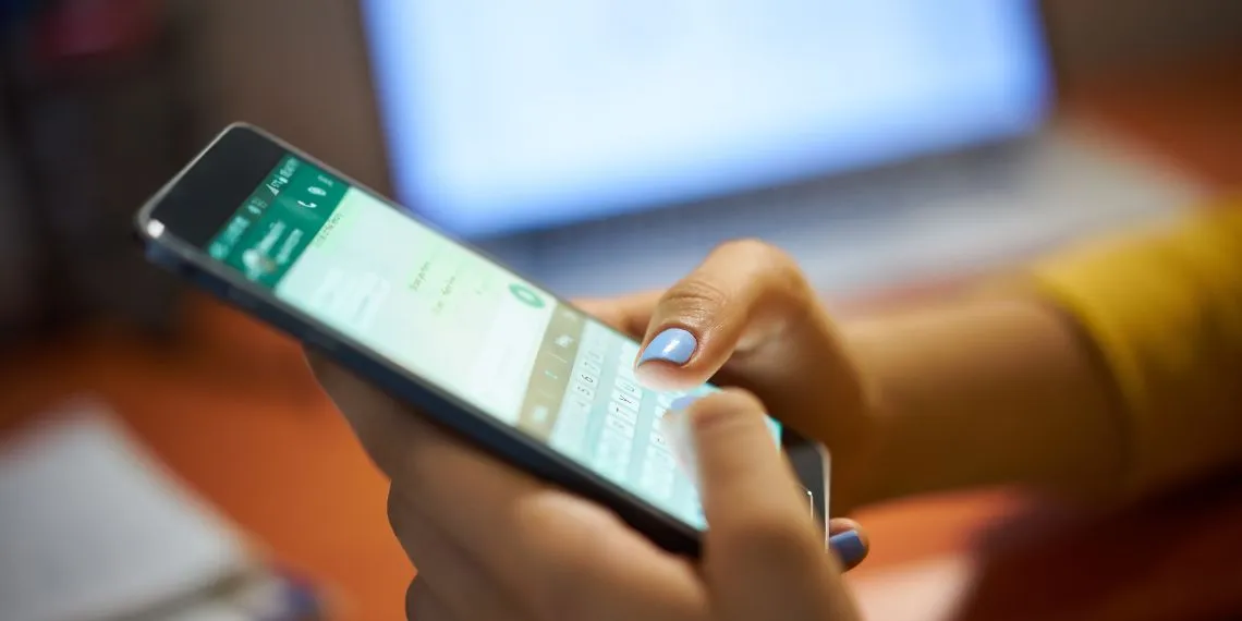 Pessoa digitando no WhatsApp pelo celular, em ambiente de trabalho com notebook ao fundo, representando o uso do aplicativo para ganhar dinheiro online.