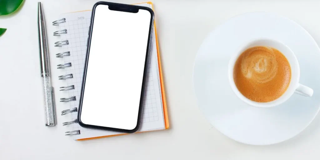 Celular sobre um caderno com grade quadriculada, ao lado de uma caneta prateada e uma xícara de café em uma escrivaninha branca, representando um kit de trabalho minimalista para produtividade em casa